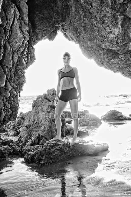 Jeune femme, triathlète à l'entraînement en bonnet de bain et lunettes vues à travers une arche de roche sur une plage — Photo de stock