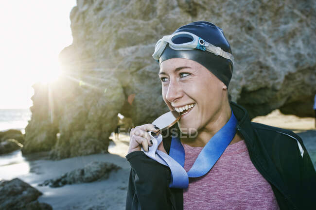 Mujer joven, triatleta con chaqueta mordiendo una gran medalla con sus dientes, una ganadora - foto de stock