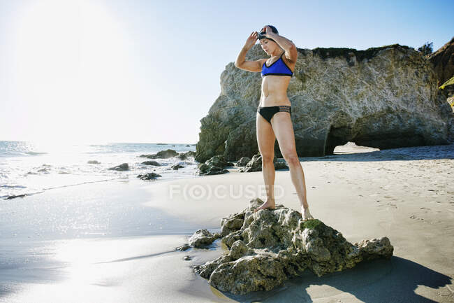Young woman, triathlete in training in swimwear, swim hat and goggles on a beach. — Stock Photo