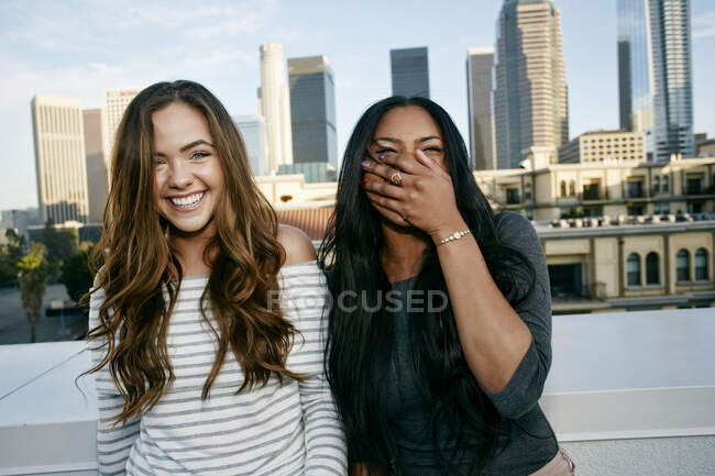 Two young women on a rooftop posing for photographs, city skyline background. — Stock Photo