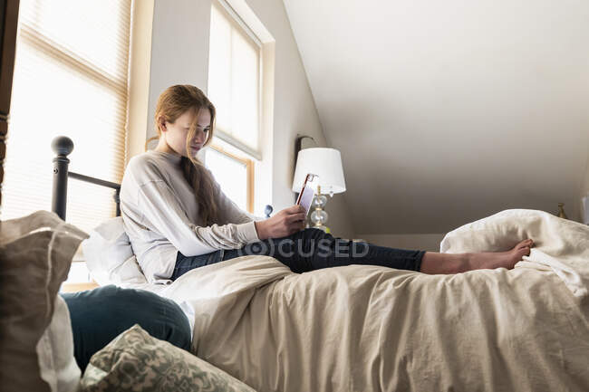 Дівчина-підліток сидить на ліжку, використовуючи свій смартфон — стокове фото