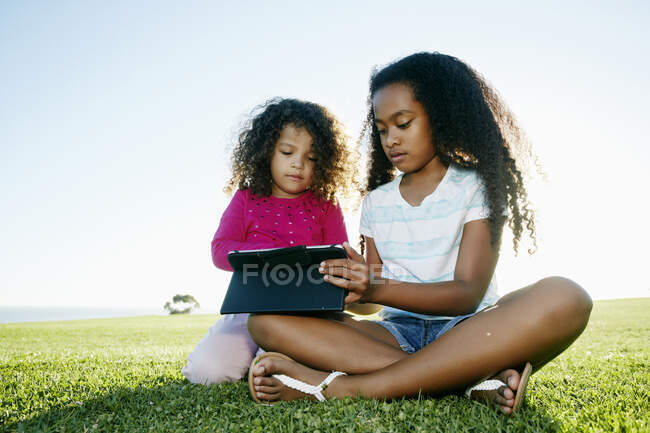 Giovane ragazza di razza mista e sua sorella minore seduti all'aperto condividendo un tablet digitale — Foto stock