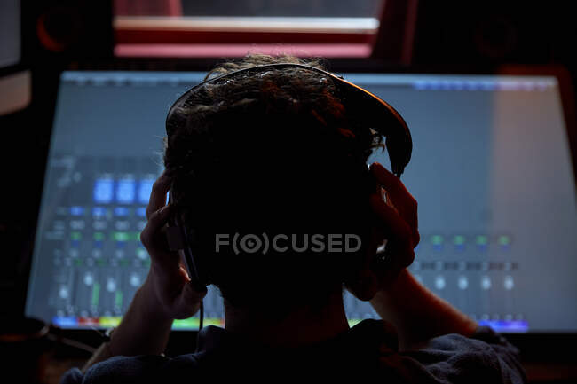 Человек, работающий в музыкальной студии в наушниках, используя большой экран компьютера, снятый сзади — стоковое фото