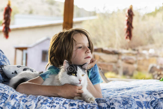 Young boy lying on outdoor bed embracing cat — Photo de stock