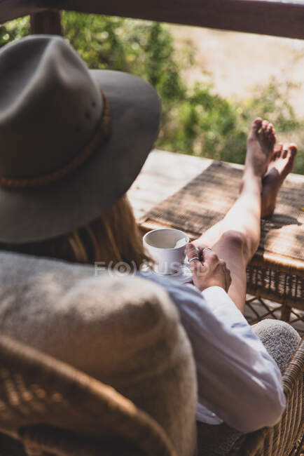 Woman sitting with her feet up drinking a cup of tea, wearing a safari hat — Stock Photo