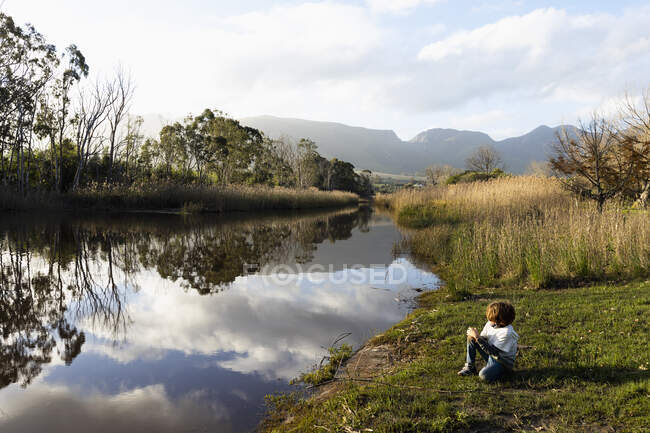 Giovane ragazzo che gioca su una riva del fiume, acqua calma piatta e spazi aperti — Foto stock