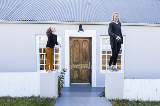 Брат і сестра стоять на низькій стіні біля будинку, сміючись — стокове фото