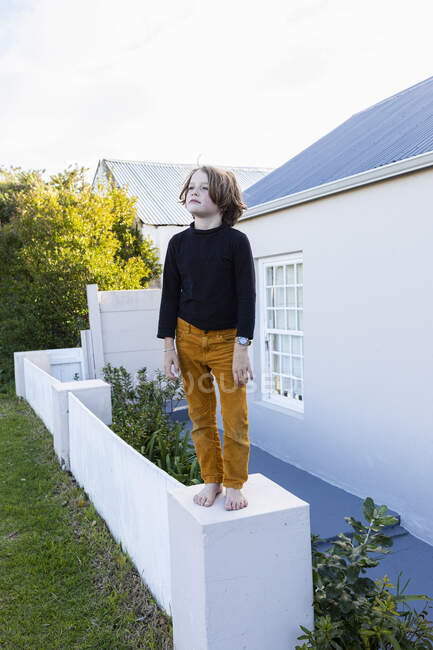 Мальчик, стоящий босиком на низкой стене возле дома — стоковое фото