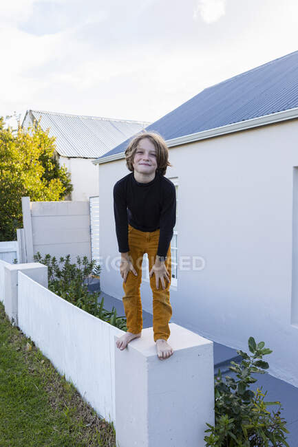 Um menino de 8 anos em pé em uma parede fora de uma casa, posando para a câmera. — Fotografia de Stock