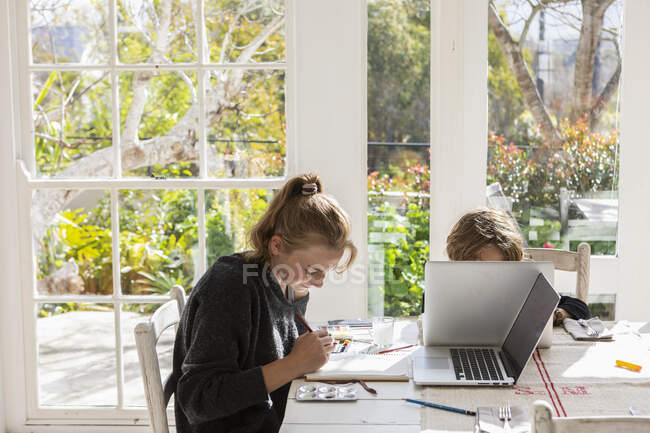 Une adolescente peint avec des aquarelles à une table, et un garçon sur un ordinateur portable — Photo de stock