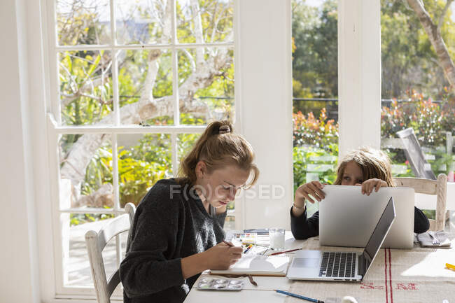 Adolescente pintando com aquarelas em uma mesa, e um menino em um laptop — Fotografia de Stock