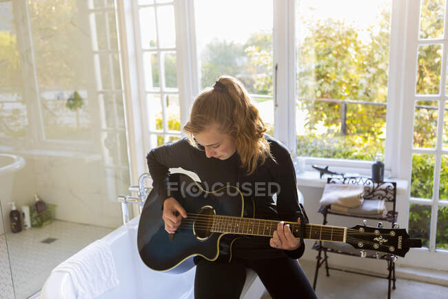 Adolescente sentada na beira de uma banheira, tocando guitarra acústica. — Fotografia de Stock