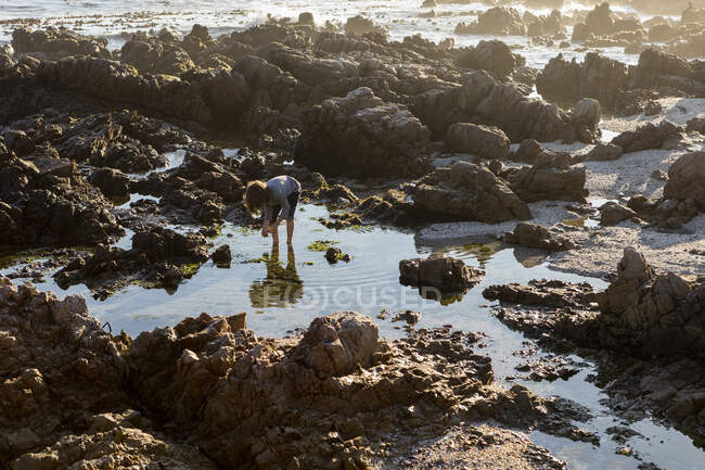 Jovem explorando uma piscina de rocha entre as rochas irregulares da costa do Oceano Atlântico ao pôr do sol — Fotografia de Stock