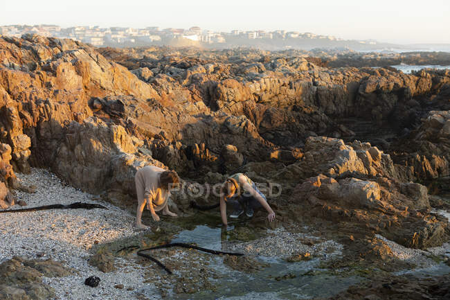 Teenage girl and her brother bending down over a rock pool among jagged rocks at sunset, De Kelders, Western Cape, South Africa. — Stock Photo