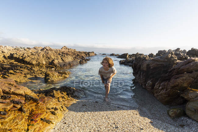 Um jovem rapaz em águas pouco profundas entre rochas irregulares na praia de De Kelders. — Fotografia de Stock