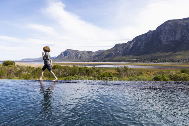 Восьмилетний мальчик прогуливается по краю бесконечного бассейна, на фоне гор — стоковое фото