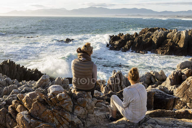 Женщина и девочка-подросток сидят на скалистом берегу и смотрят в море. — стоковое фото