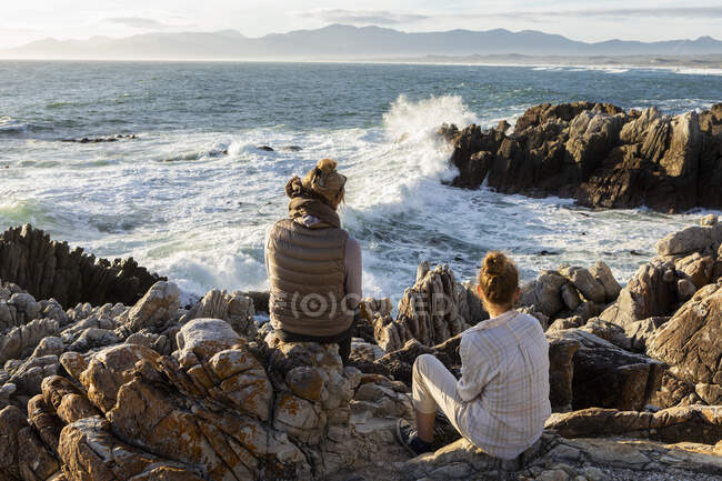 Mujer y adolescente sentado en la orilla rocosa, mirando hacia el mar. - foto de stock