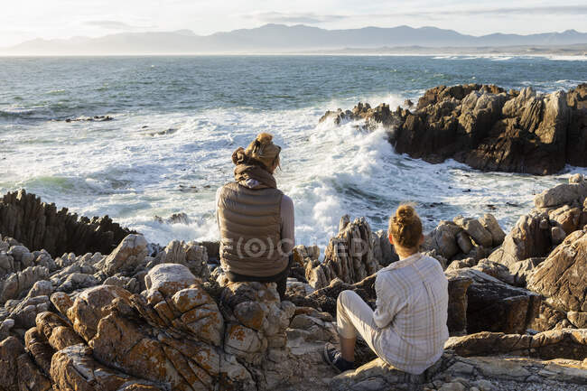Женщина и девочка-подросток сидят на скалистом берегу и смотрят в море. — стоковое фото