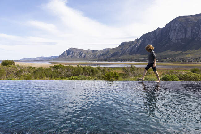 Garçon de huit ans marchant au bord d'une piscine à débordement, toile de fond de montagne — Photo de stock
