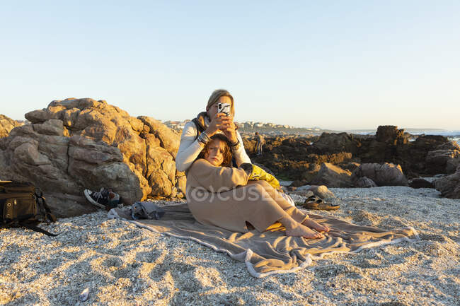 Donna e suo figlio seduti sulla sabbia tra le rocce sulla riva a De Kelders al tramonto. — Foto stock