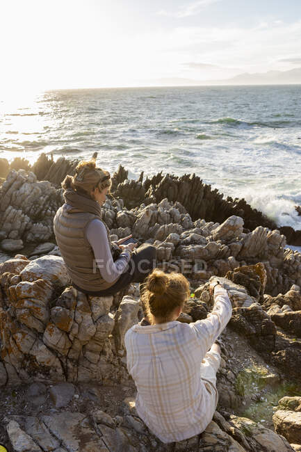 Woman and teenage girl sitting on rocky shore, looking out to sea. — Stock Photo