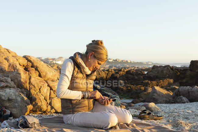 Взрослая женщина, сидящая на пляже со своим смартфоном в Де Келдерс на закате. — стоковое фото