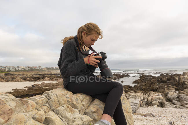 Дівчина-підліток використовує цифрову камеру, переглядаючи зображення, сидячи на каменях на пляжі . — стокове фото