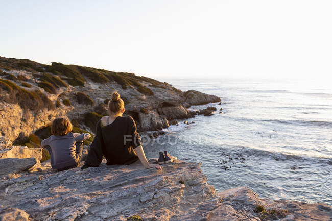 Adolescente e jovem irmão ao pôr do sol, sentados lado a lado olhando sobre o oceano. — Fotografia de Stock