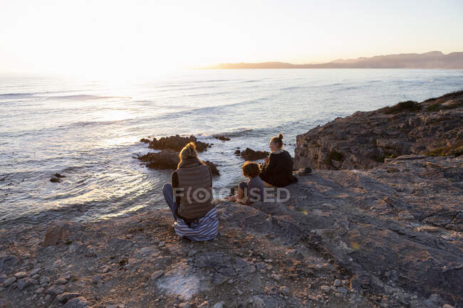 Una famiglia, una madre e due bambini seduti a guardare il tramonto sull'oceano. — Foto stock