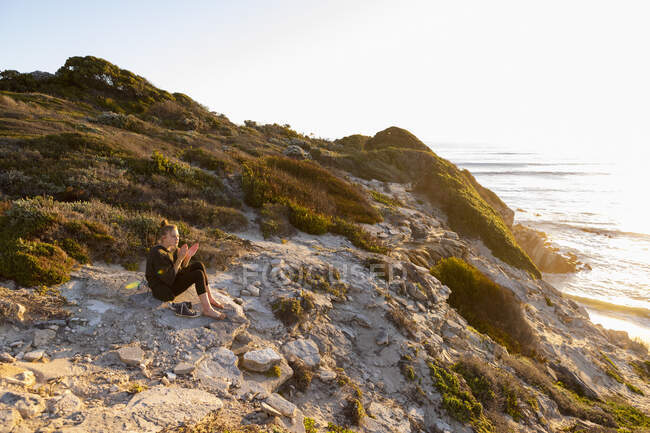 Adolescente sentada e assistindo o pôr do sol sobre a água de falésias. — Fotografia de Stock