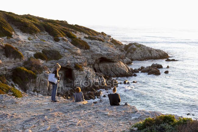 Drei Personen, eine Familie sitzt und beobachtet den Sonnenuntergang über dem Ozean. — Stockfoto