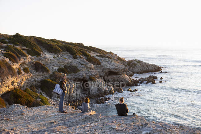 Tres personas, una familia sentada mirando la puesta de sol sobre el océano. - foto de stock