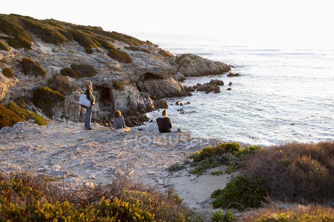 Drei Personen, eine Familie sitzt und beobachtet den Sonnenuntergang über dem Ozean. — Stockfoto