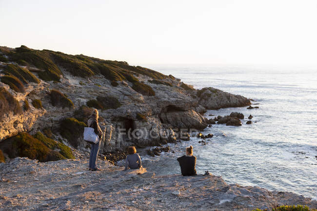 Tres personas, una familia sentada mirando la puesta de sol sobre el océano. - foto de stock