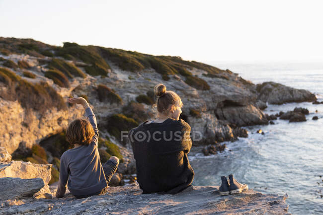 Adolescente e menino sentado em rochas olhando sobre o mar ao pôr do sol — Fotografia de Stock