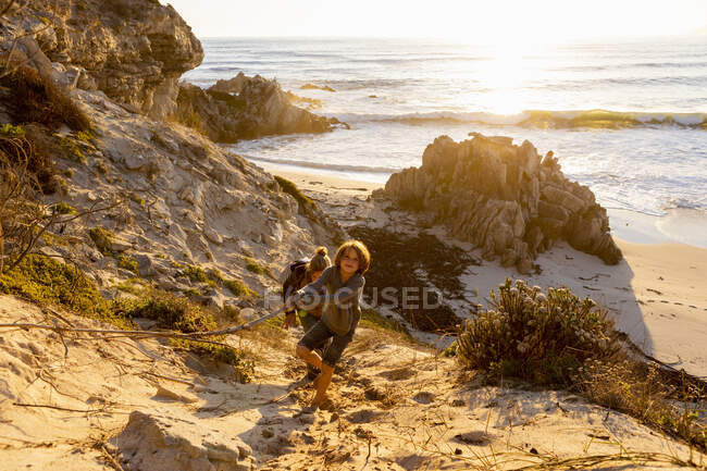 A boy and a woman clambering up a very steep sandy slope above a beach — Stock Photo