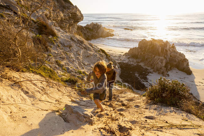 A boy and a woman clambering up a very steep sandy slope above a beach — Stock Photo