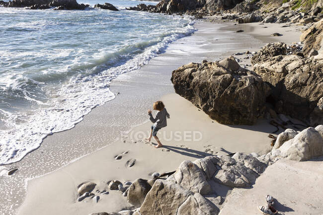 Un giovane ragazzo solo su una piccola distesa di sabbia sotto le scogliere vicino all'oceano. — Foto stock
