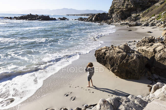 Un niño solo en un pequeño tramo de arena bajo los acantilados junto al océano. - foto de stock