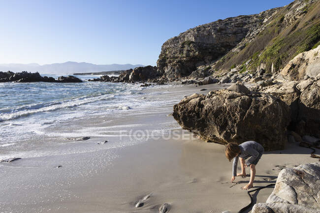 Um rapaz sozinho num pequeno pedaço de areia debaixo das falésias junto ao oceano.. — Fotografia de Stock