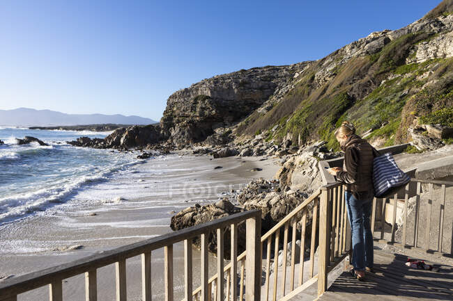 Uma mulher madura em pé em uma passarela com vista para uma praia de areia — Fotografia de Stock