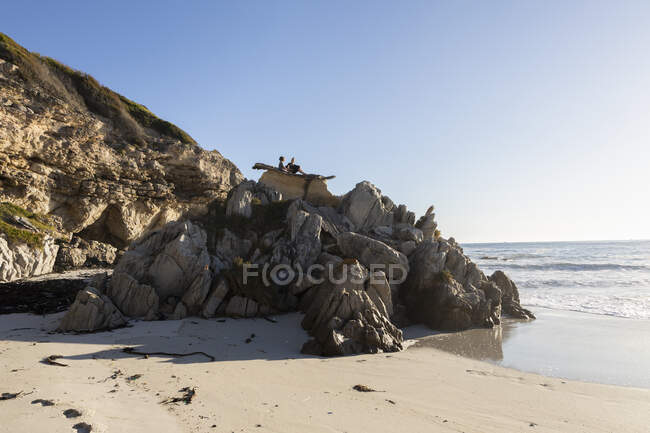 Two children perched on top of jagged rocks overlooking a sandy beach at low tide — Stock Photo