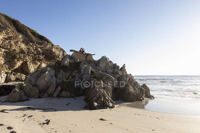 Two children perched on top of jagged rocks overlooking a sandy beach at low tide — Stock Photo