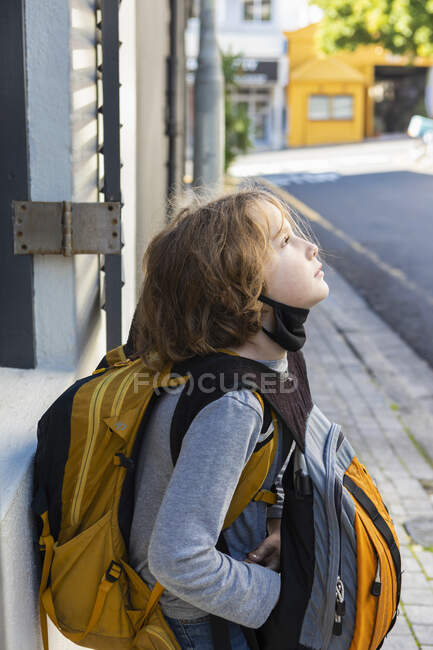 A boy with a black facemask tucked under his chin, on a street with a backpack and bag. — Stock Photo