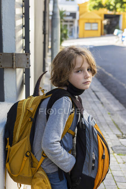 A boy with a black facemask tucked under his chin, on a street with a backpack and bag. — Stock Photo