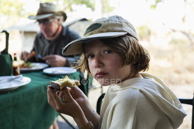 Giovane ragazzo mangiare un pezzo di pane tostato a un tavolo in un campo di safari — Foto stock