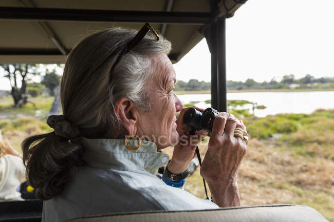 Donna anziana con binocolo, seduta in un veicolo safari, che guarda oltre paludi e corsi d'acqua — Foto stock