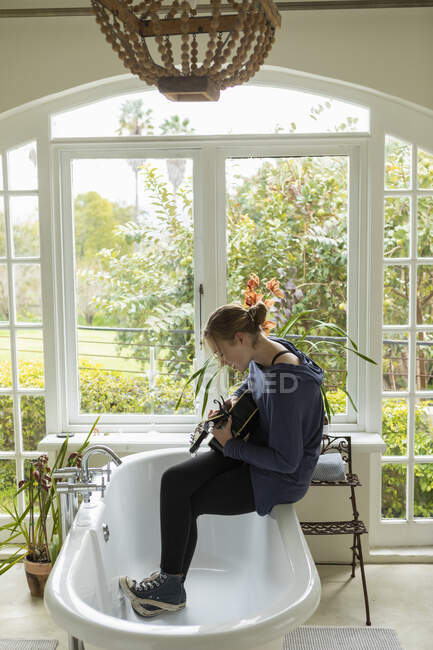 Teenage girl sitting on the edge of a bathtub playing guitar and singing. — Stock Photo