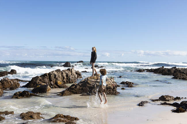 Duas crianças explorando em uma praia de areia com rochas irregulares e ondas quebrando, — Fotografia de Stock